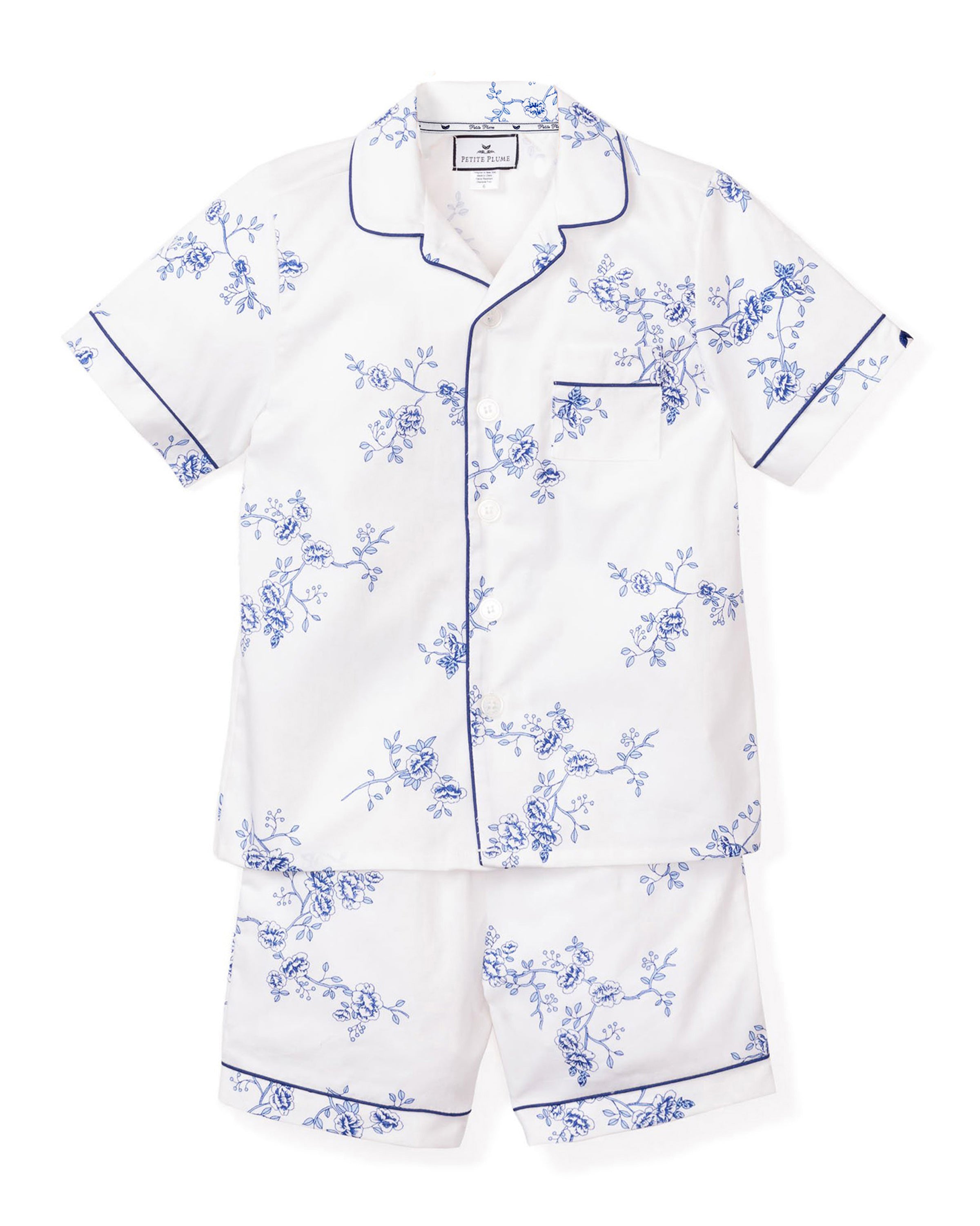 The Kids Twill Pajama Short Set in Indigo Floral by Petite Plume includes white cotton pajamas with a blue floral print. The top has short sleeves, a collar, chest pocket, and blue piping. The shorts feature an elastic waistband and matching piping for luxurious comfort.