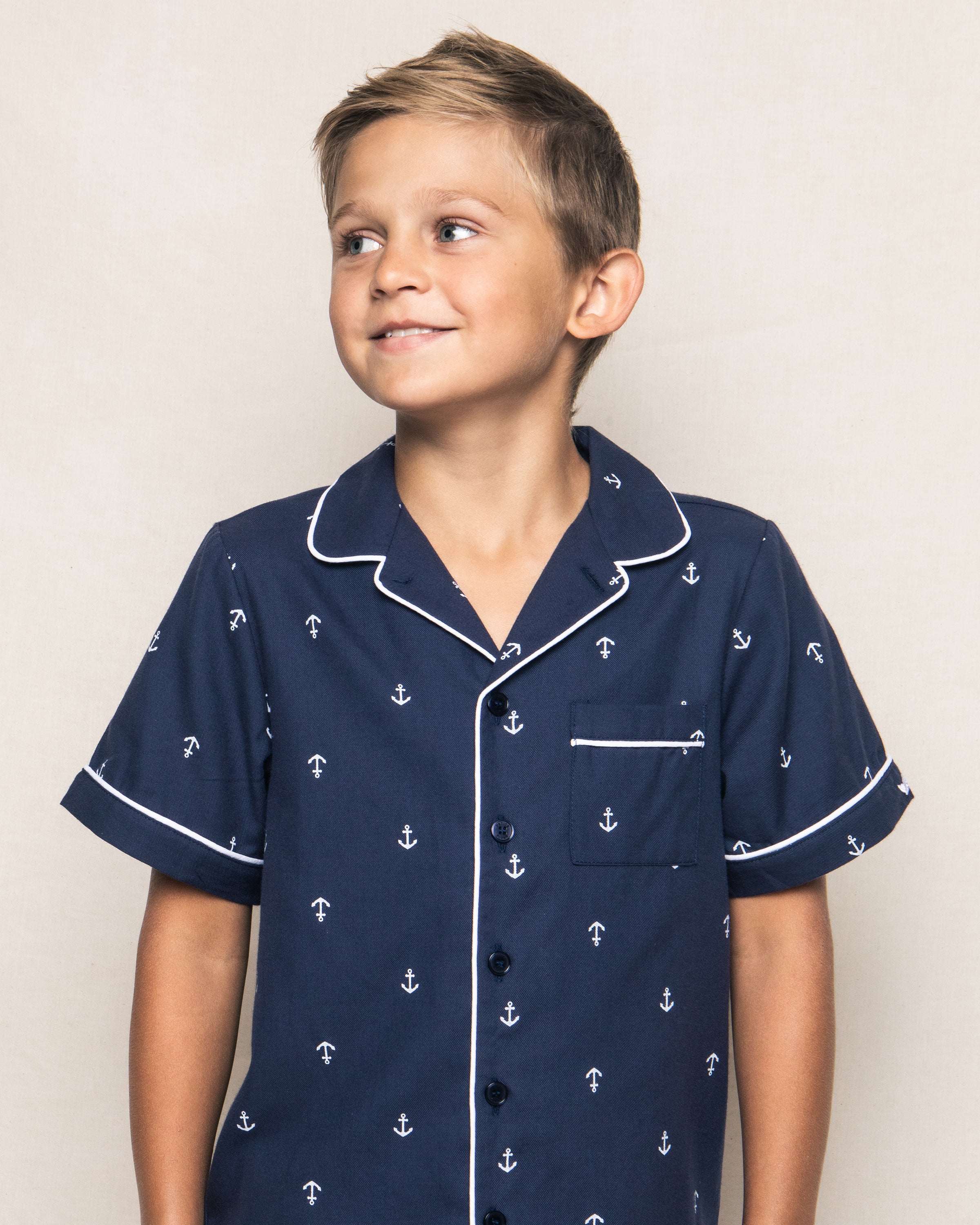 A young boy smiles to the side wearing Petite Plumes navy blue Kids Twill Pajama Short Set in Portsmouth Anchors, adorned with small white anchors. The shirt features white piping around the collar and pocket, evoking a luxurious sleepwear feel against a neutral background.