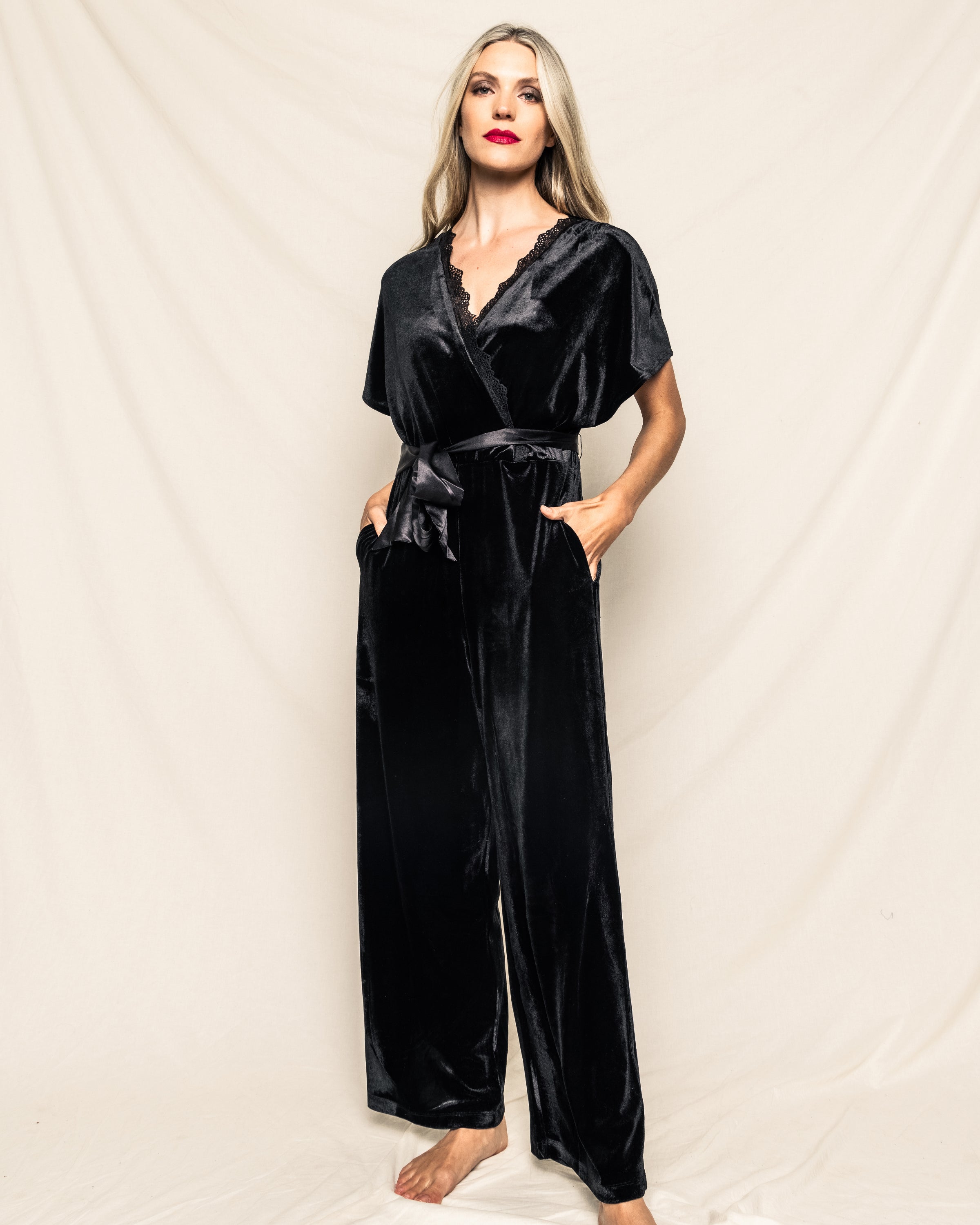 Against a light background, a long-haired person exudes Hollywood glamour in Petite Plumes Womens Velour Jumpsuit in Black. With one hand in a pocket, short sleeves, waist tie, and bright red lipstick, they stand barefoot radiating classic elegance.
