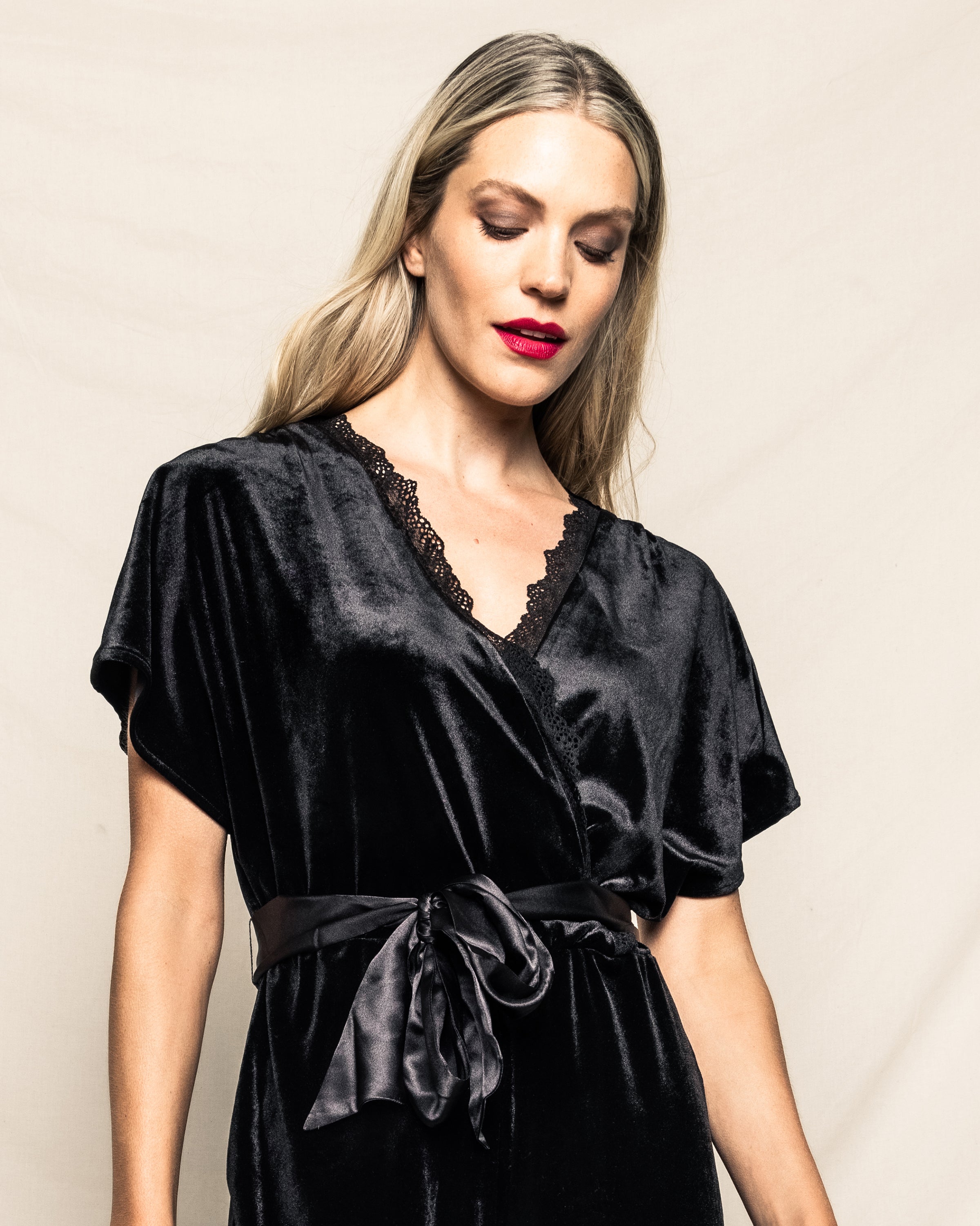 A woman with long blonde hair exudes glamour in a Petite Plume Womens Velour Jumpsuit in Black, posing against a neutral background. Her red lipstick and subtle, confident expression channel classic Hollywood style.
