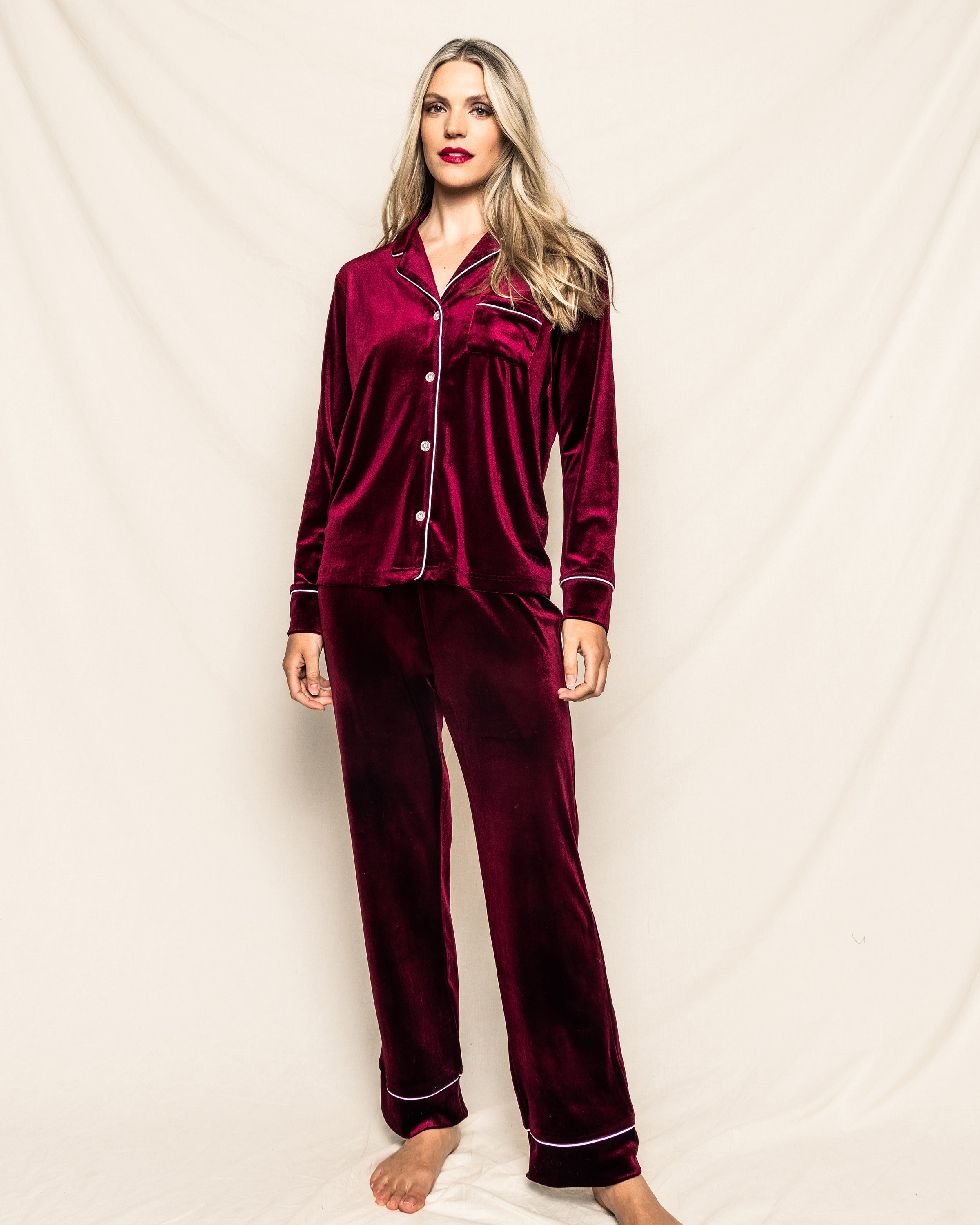 A person with long hair stands against a cream backdrop, wearing the Womens Velour Pajama Set in Royal Garnet by Petite Plume. With white piping evoking classic Hollywood style from the Cabaret Collection, they maintain a relaxed posture while looking at the camera.