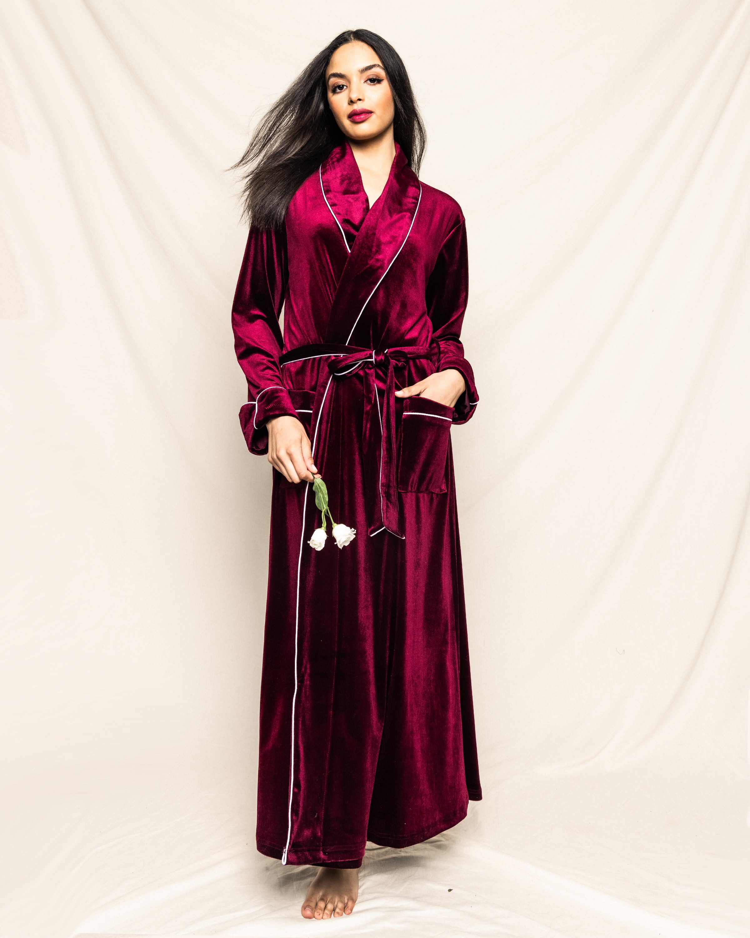 A person in Petite Plumes Womens Velour Robe in Royal Garnet stands against a light draped background, holding two white tulips and facing forward. This Hollywood-style robe from the Cabaret Collection has a belt and large front pockets, epitomizing elegant sleepwear.