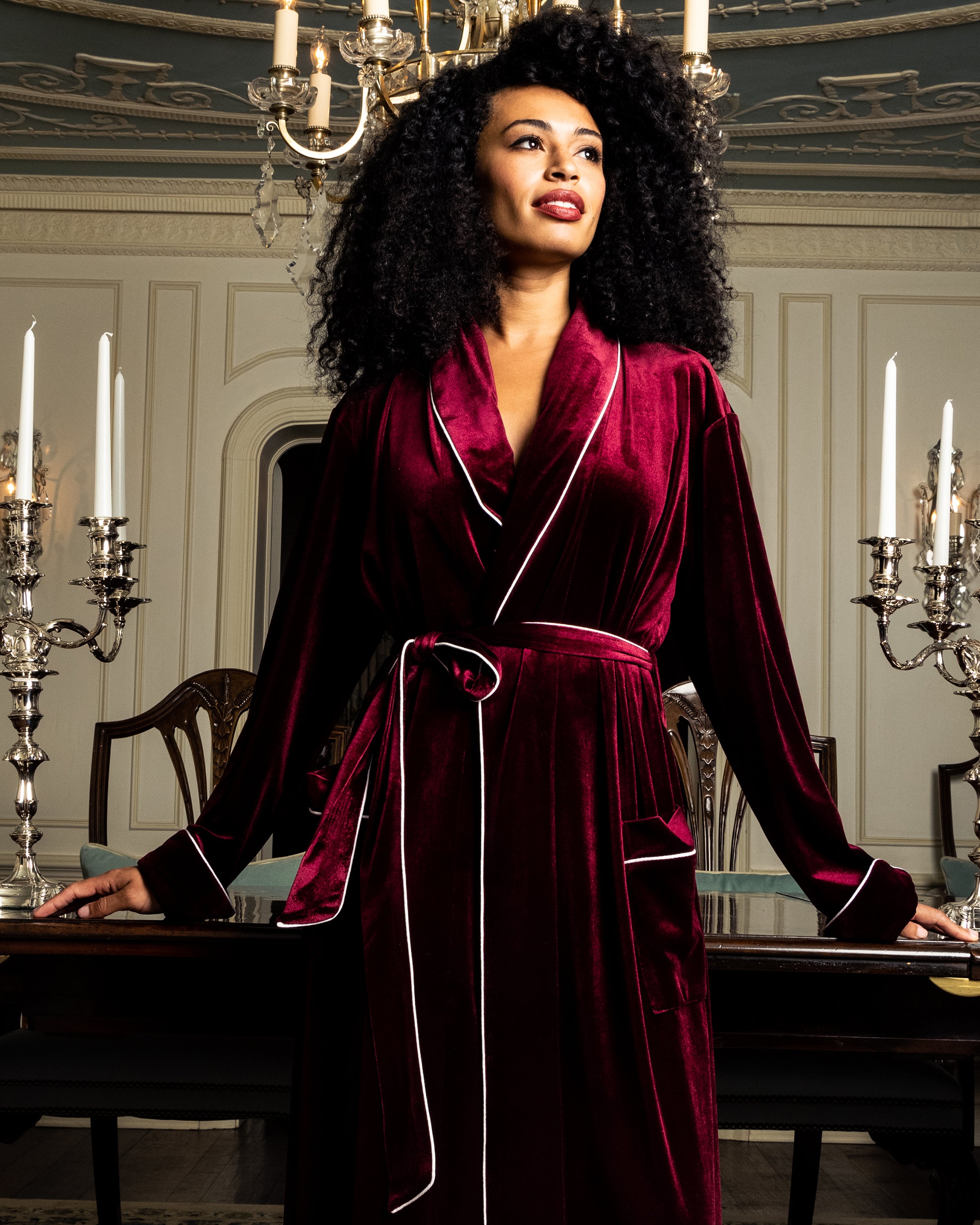 A woman with curly hair confidently stands in an elegant room wearing the Womens Velour Robe in Royal Garnet by Petite Plume. The space boasts a chandelier, tall candles, and ornate wall designs, creating a luxurious Hollywood style atmosphere.