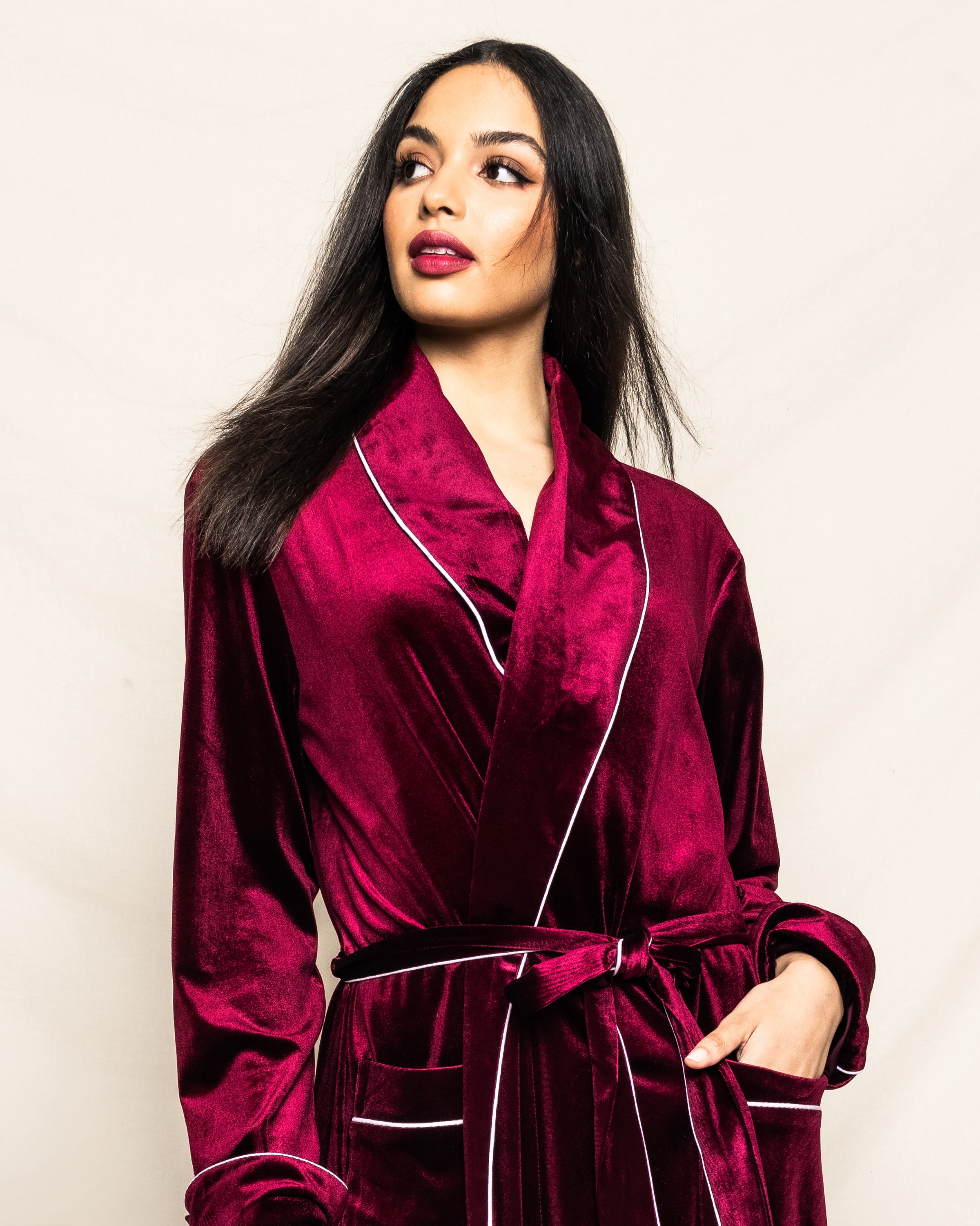 A woman with long dark hair wears a Royal Garnet velour robe with white piping from Petite Plume, embodying the exquisite sleepwear of the Cabaret Collection. She poses against a light background, looking to the side.