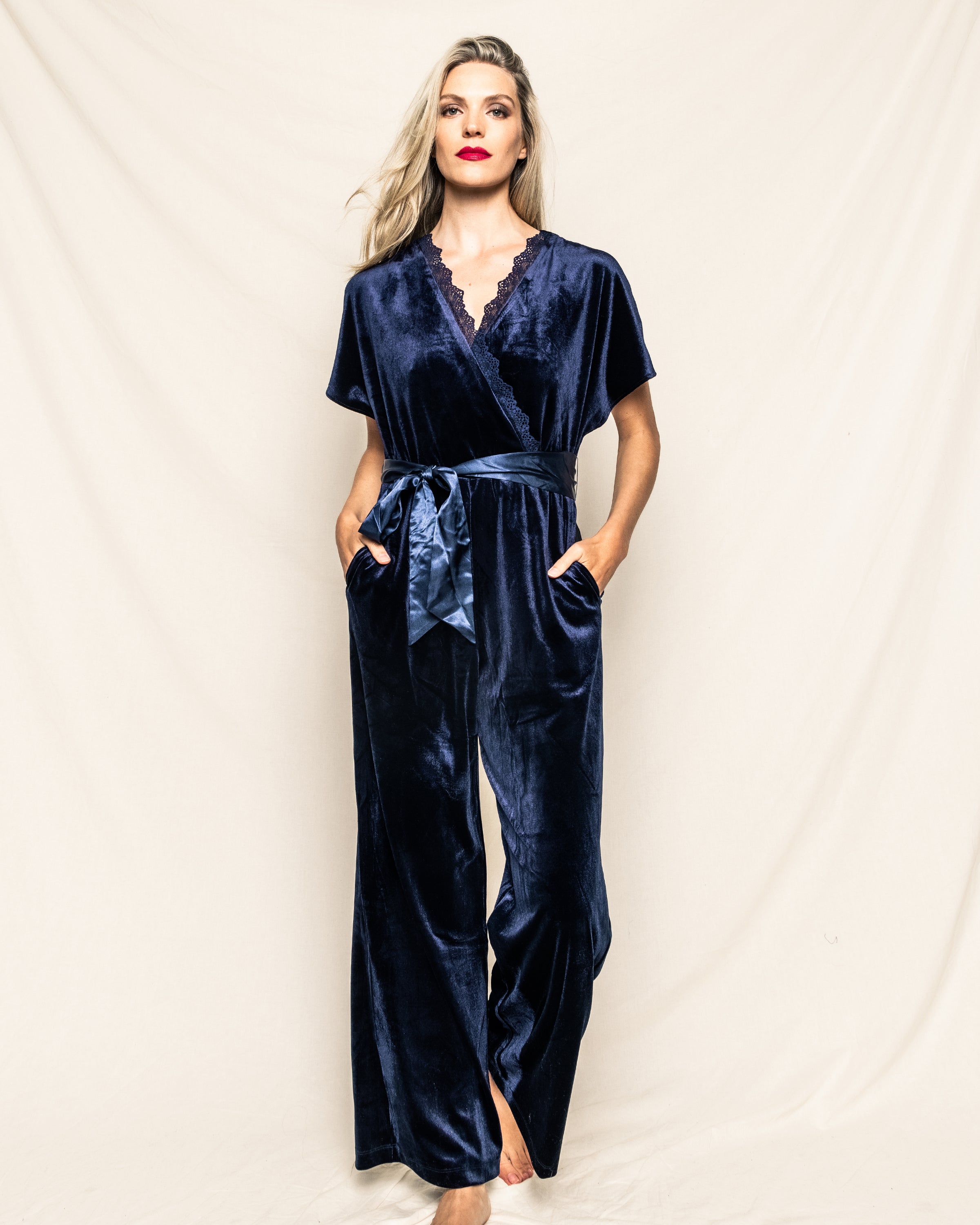 Wearing Petite Plumes Womens Velour Jumpsuit in Navy, a person stands barefoot with confidence against a cream backdrop. The lace-trimmed neckline and satin tie belt add to the classic Hollywood style, as they rest their hands in their pockets.