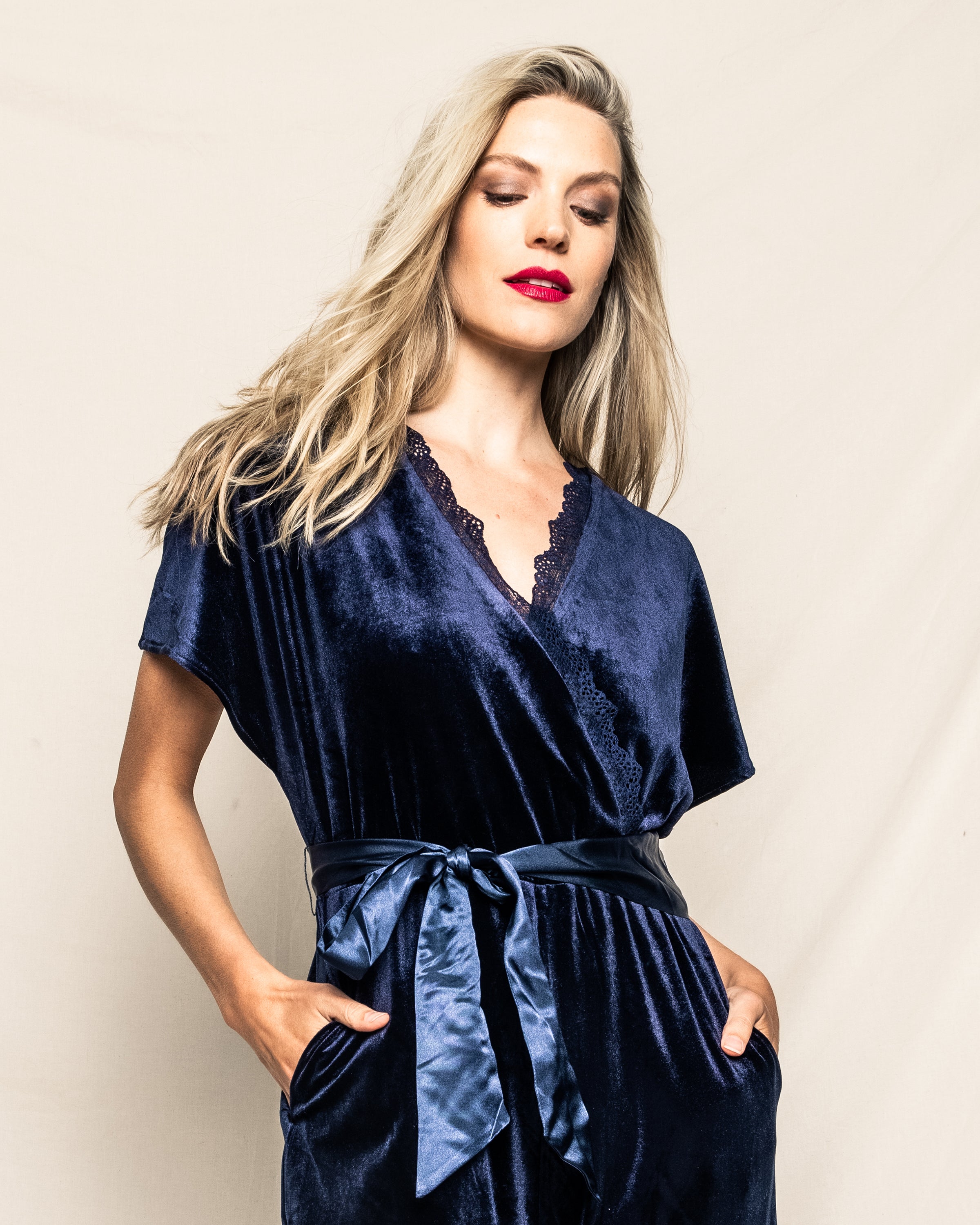 A person with long blonde hair and red lipstick poses confidently in the Womens Velour Jumpsuit in Navy by Petite Plume. The lace trim and matching satin belt accentuate its classic Hollywood style, with hands casually resting in pockets against a plain background.