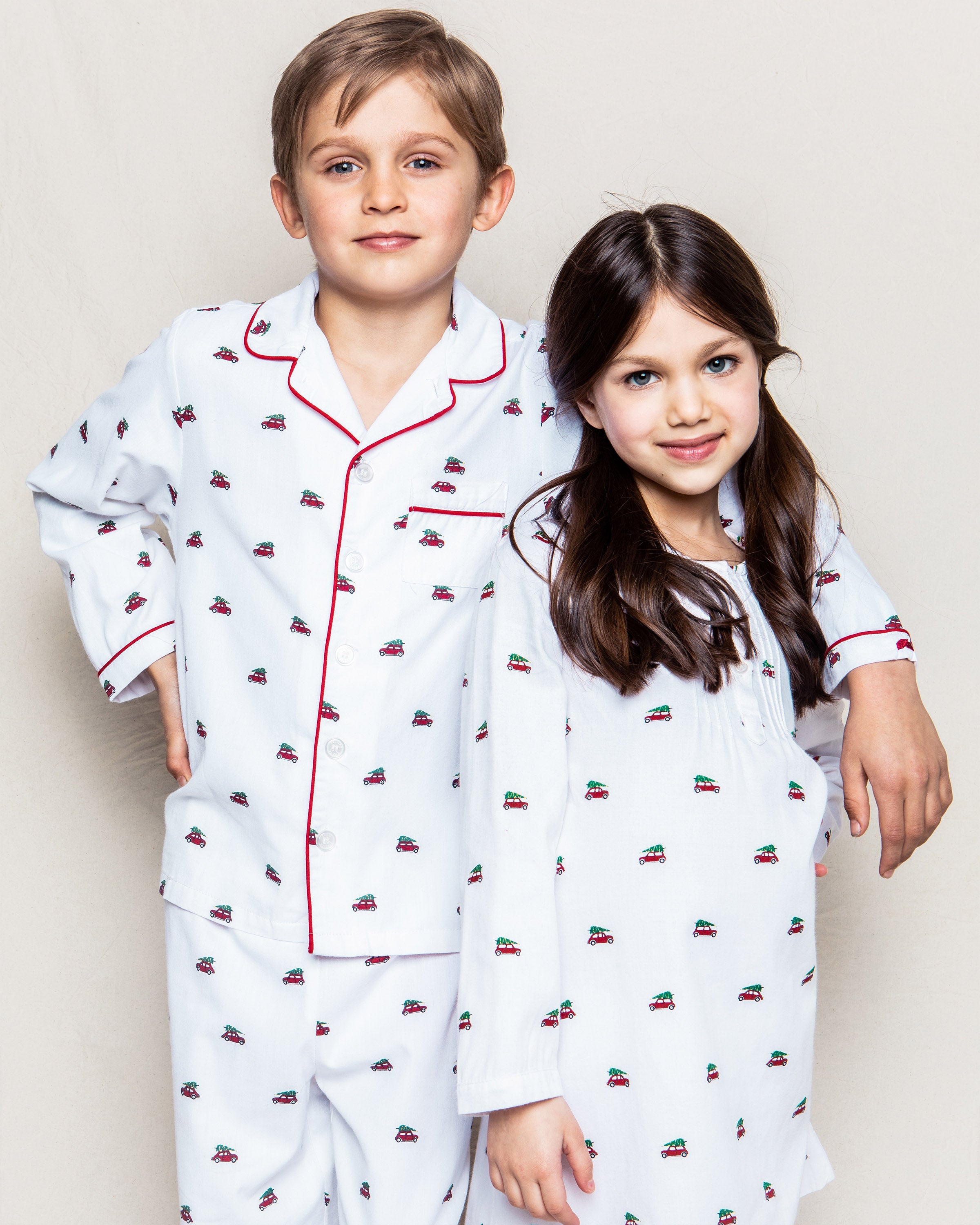 Two children in luxury sleepwear display matching Petite Plume attire. The boys gentle hand rests on the girls shoulder as they both smile softly, showcasing the Girls Twill Beatrice Nightgown in Holiday Journey with small red and black patterns against a plain backdrop.