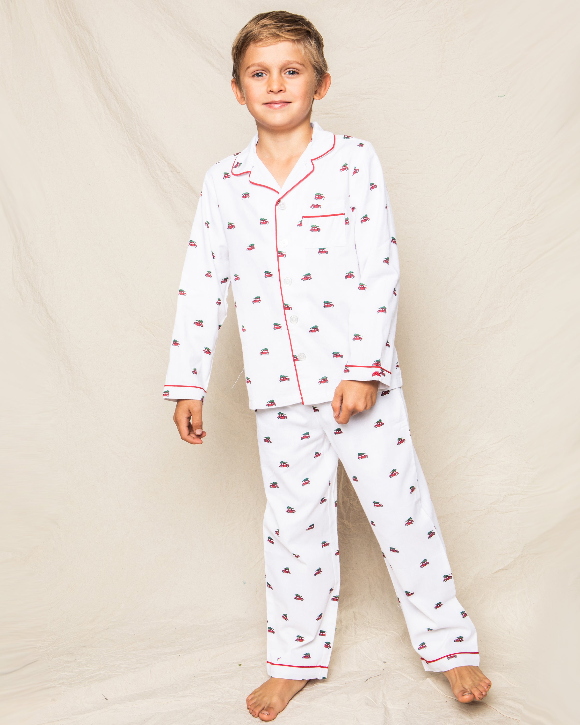 A young boy smiles barefoot against a neutral backdrop, wearing Petite Plumes cozy cotton Twill Pajama Set in Holiday Journey with red and black patterns and charming red piping on the collar and cuffs.