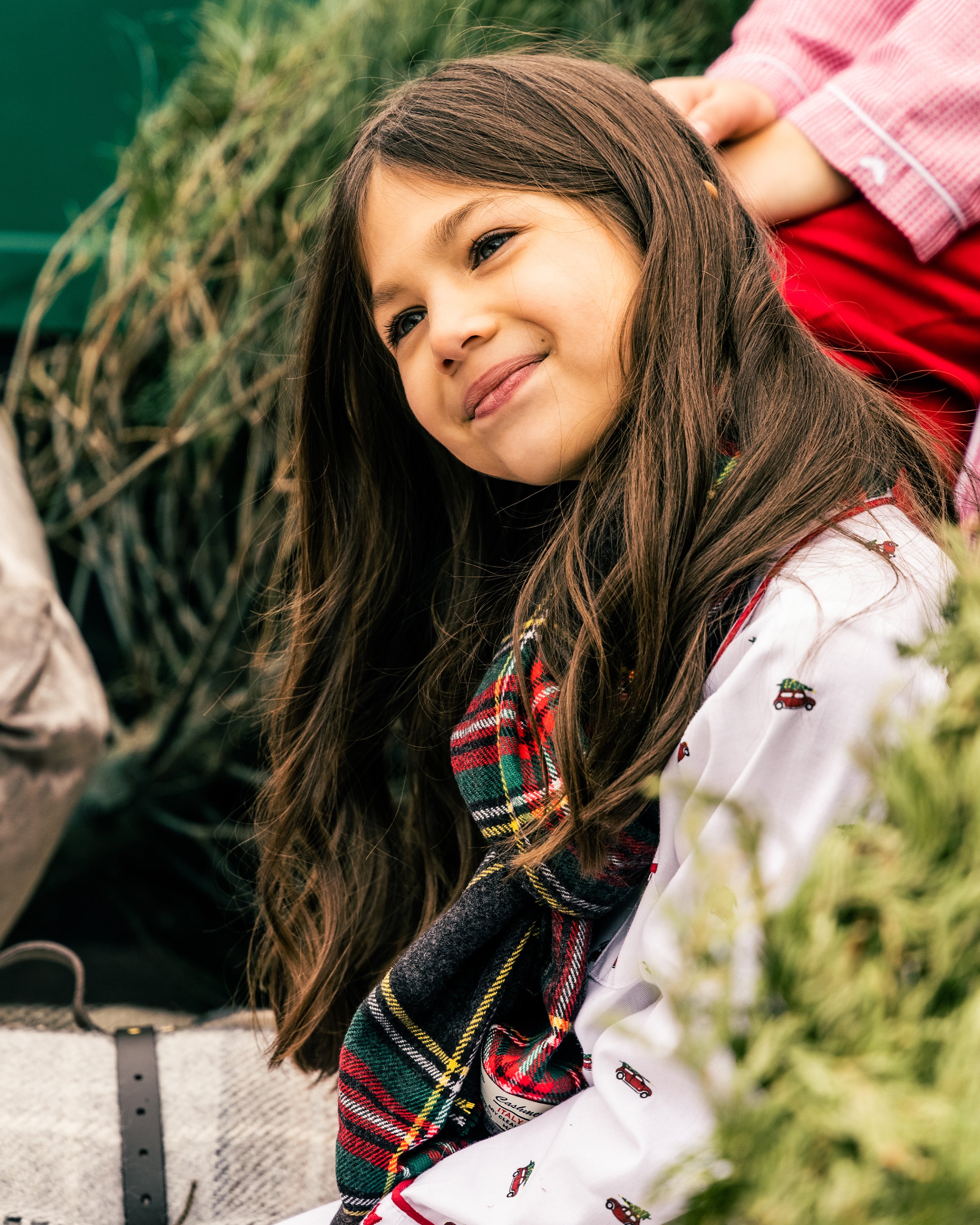 A young girl with long brown hair smiles softly while looking up. She wears Petite Plumes Kids Twill Pajama Set in Holiday Journey, featuring a white shirt with small designs and a colorful plaid scarf, perfect for her holiday journey. Greenery surrounds her, creating a peaceful setting.