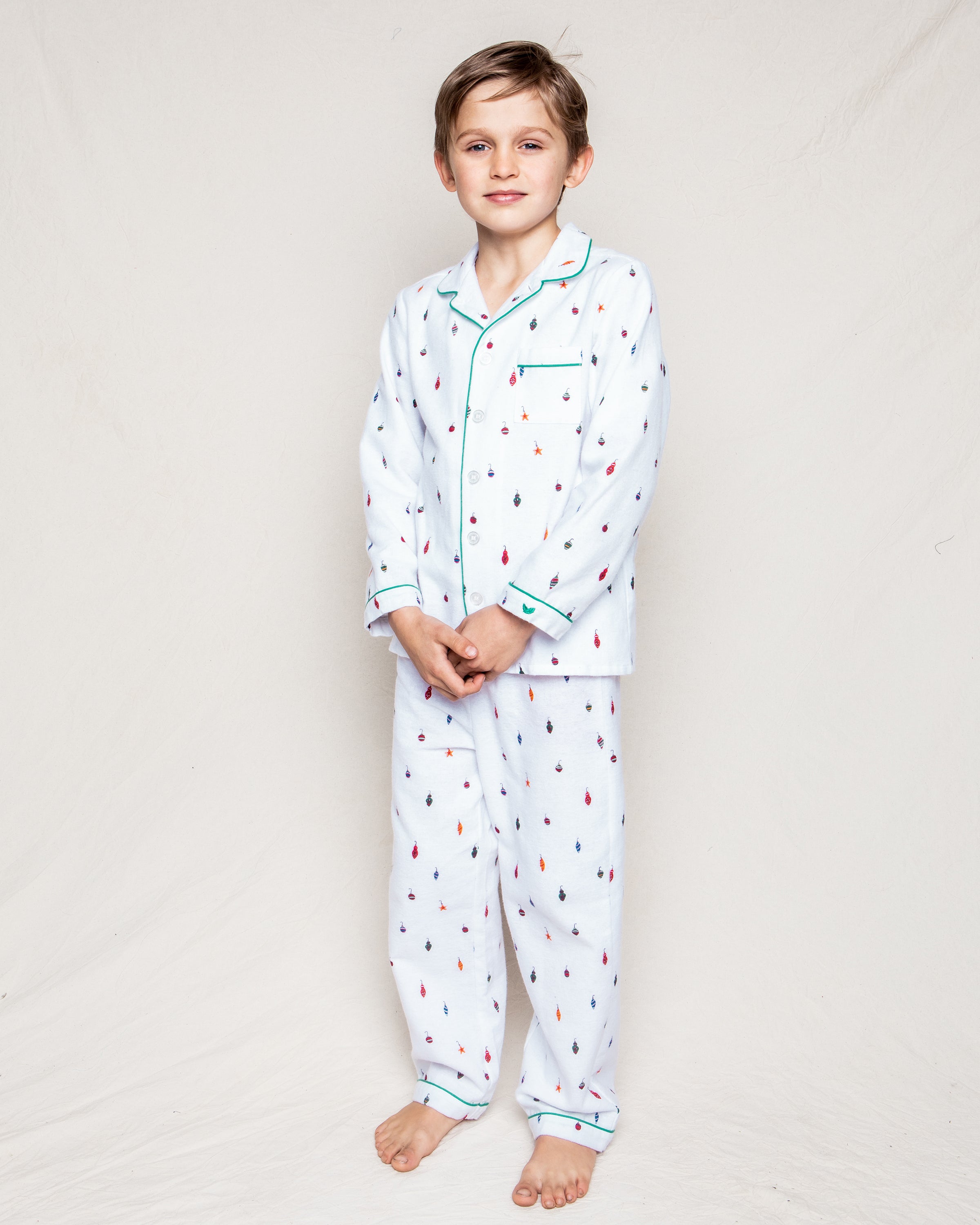 A barefoot young boy with short, light brown hair and a neutral expression stands against a plain background, wearing Petite Plumes Kids Flannel Pajama Set in Yuletide Ornaments. His hands are clasped in front of him.