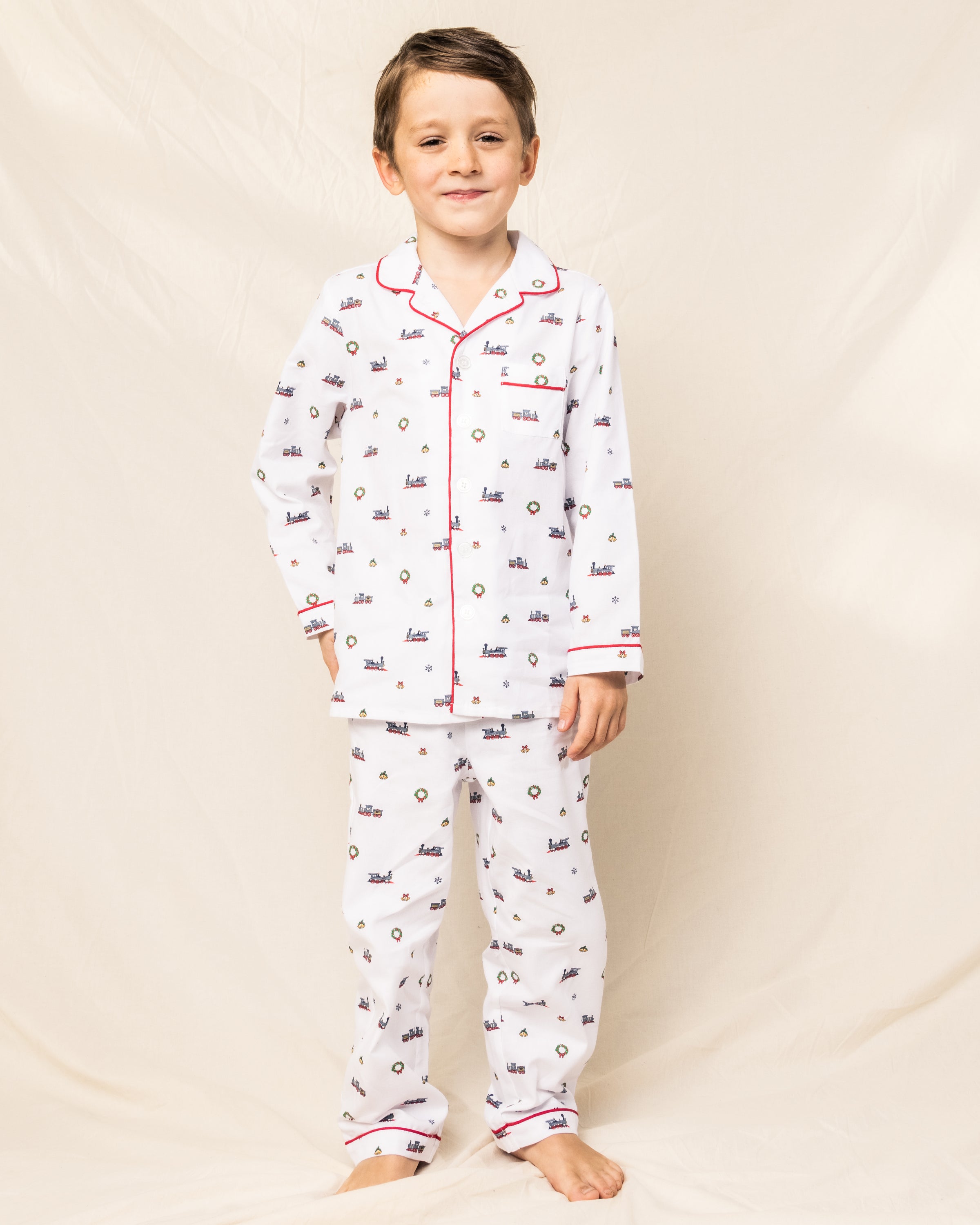 A child beams in a Petite Plume Kids Twill Pajama Set, Arctic Express edition featuring small patterns and red piping. On a soft beige background, they stand barefoot on a smooth surface, radiating warmth and comfort.