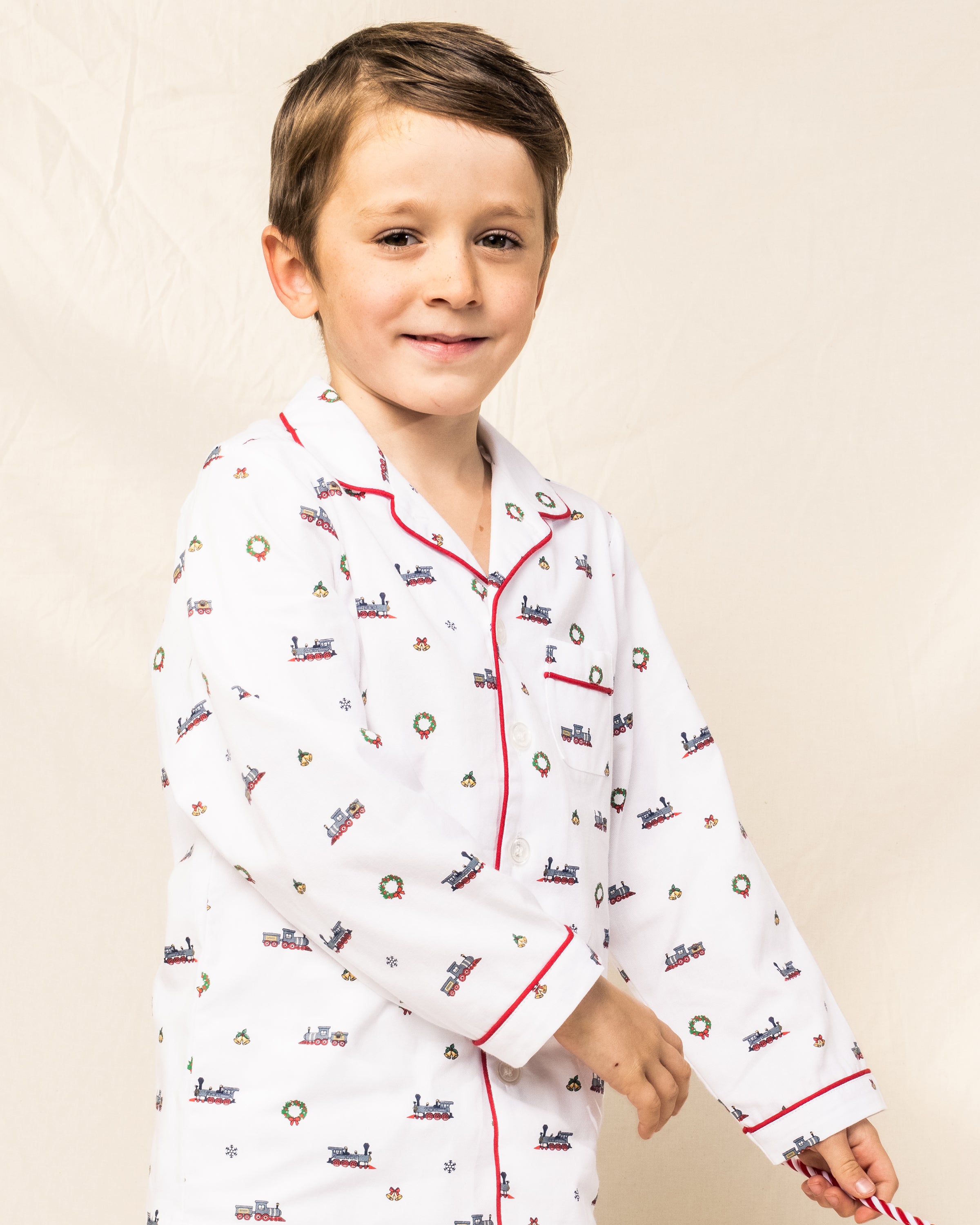 A young boy with short brown hair smiles in a Petite Plume Kids Twill Pajama Set, Arctic Express design, featuring red piping and small trains, wreaths, and snowflakes. The flame-retardant sleepwear stands out against the plain light-colored background.