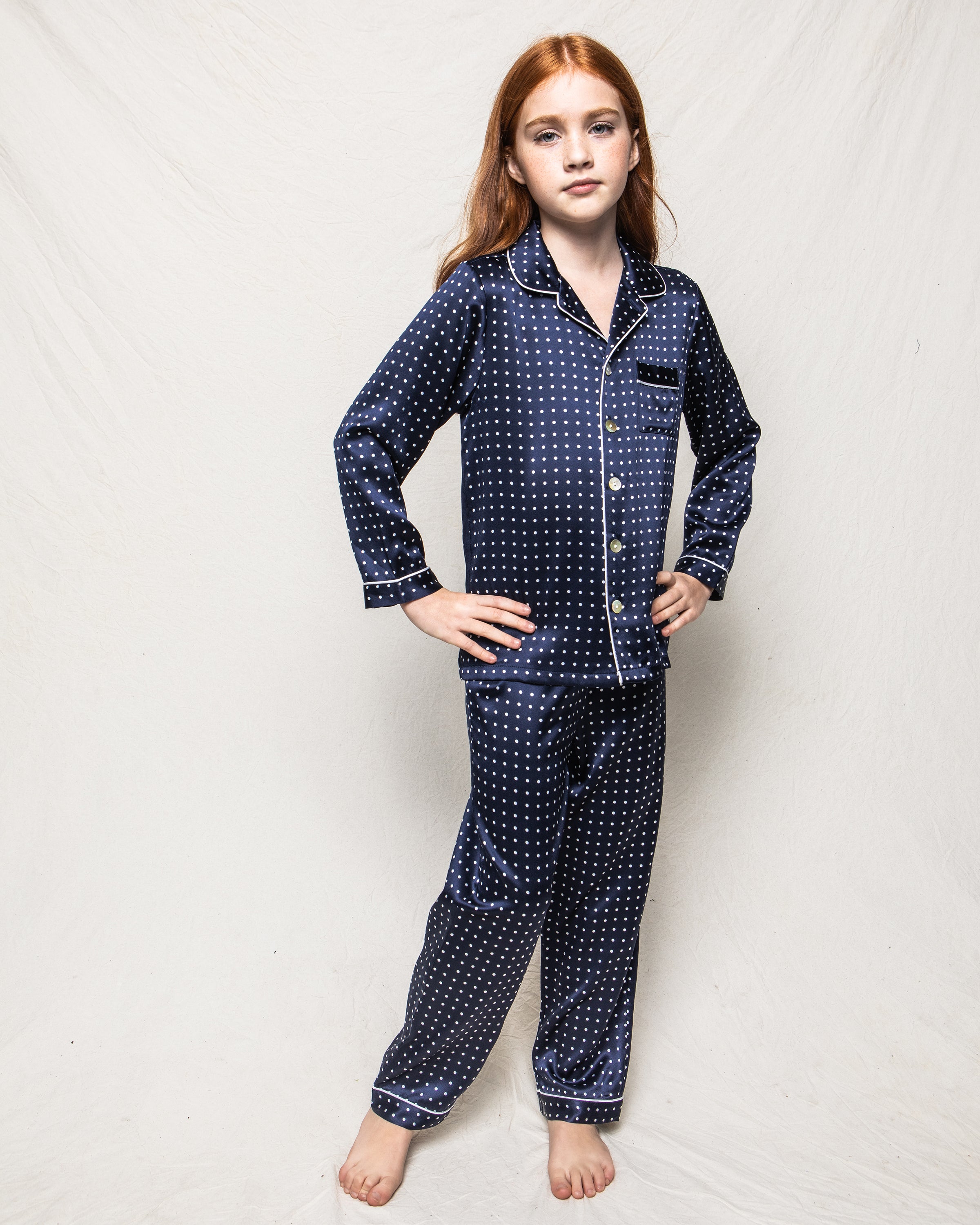 A young girl with long red hair models the Kids Silk Pajama Set in Navy Polka Dot by Petite Plume. She stands barefoot against a neutral backdrop, looking confidently at the camera.