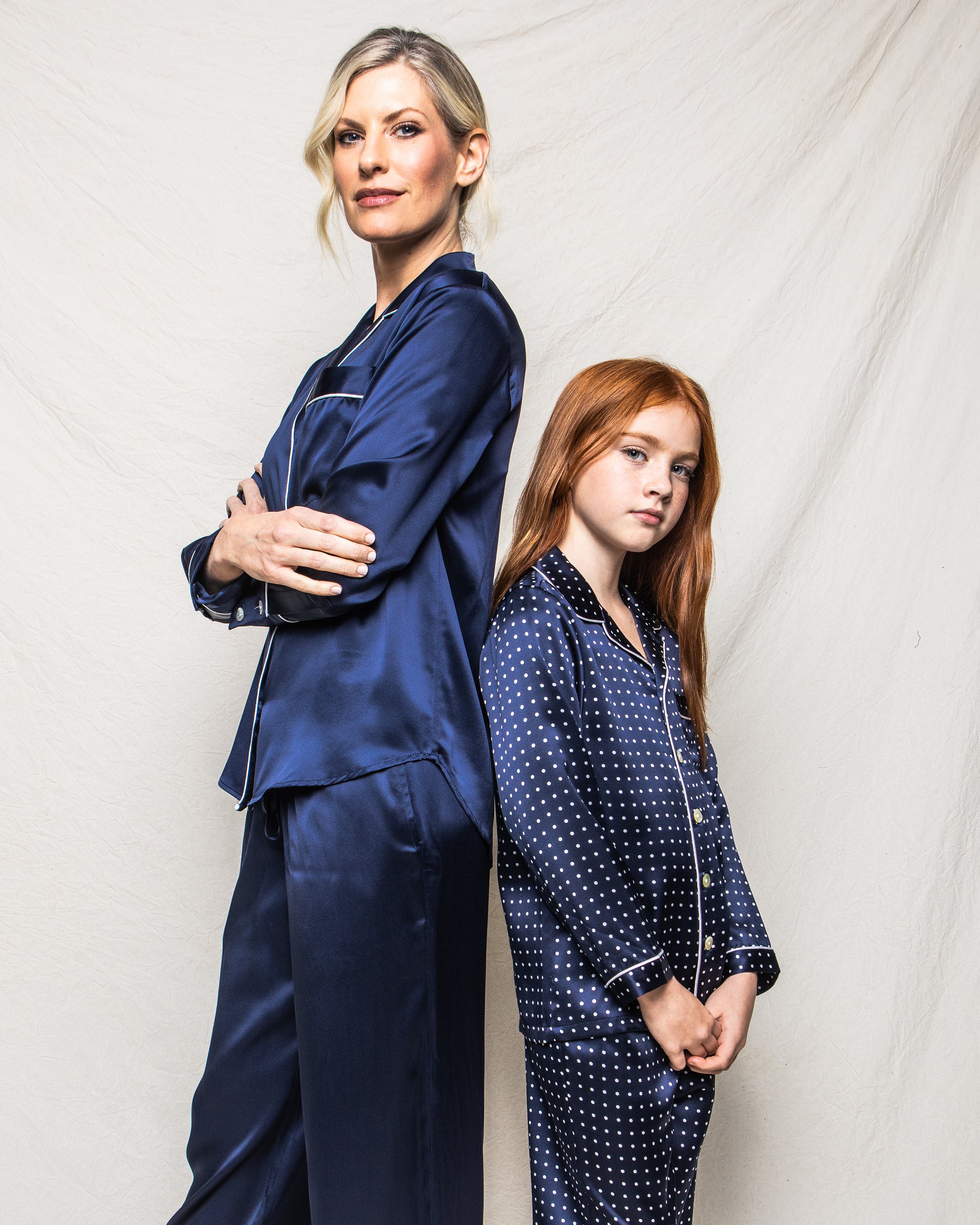 A woman and a young girl stand back-to-back in Petite Plumes silk pajamas, both adorned with navy polka dots. The woman crosses her arms confidently while the girl gazes forward, with a plain, light background that highlights their elegant Kids Silk Pajama Set in Navy Polka Dot.